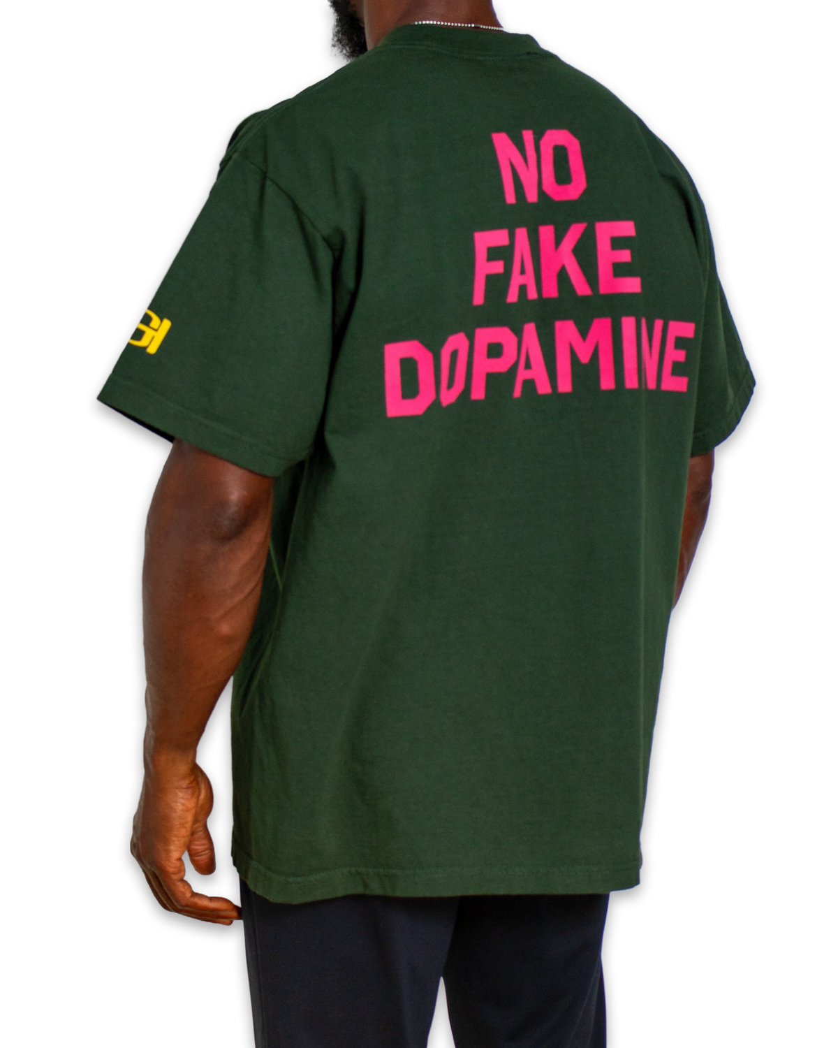 A man wearing a dark green t-shirt from the back, featuring the text 'NO FAKE DOPAMINE' printed in bright pink across the back, with a small yellow graphic on the left sleeve.