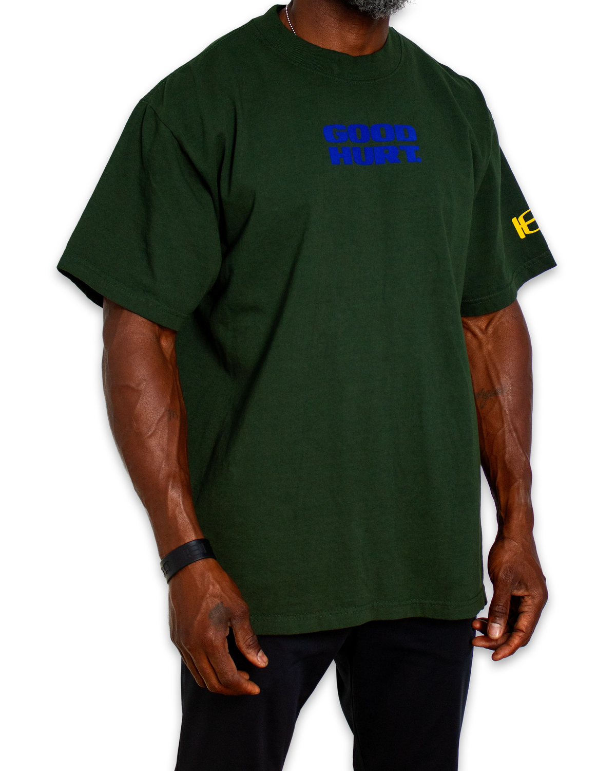 A dark green short-sleeve t-shirt with 'GOOD HURT' printed in blue on the chest and a yellow graphic on the right sleeve, shown on a model against a white background.
