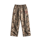 A pair of camouflage pants featuring a realistic tree bark and leaf pattern in shades of brown, beige, and gray, with a 'GOOD HURT' Athletics logo on the upper right leg.
