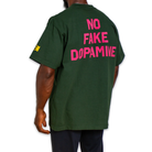 A man wearing a dark green t-shirt from the back, featuring the text 'NO FAKE DOPAMINE' printed in bright pink across the back, with a small yellow graphic on the left sleeve.